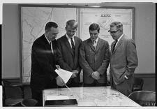 Black and white photo of four men in suits examine a tabletop map in an office setting, with additional maps displayed on the wall behind them.