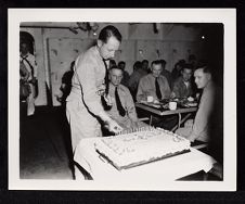 USS Saratoga birthday party, men in mess hall with one lighting candles on cake 